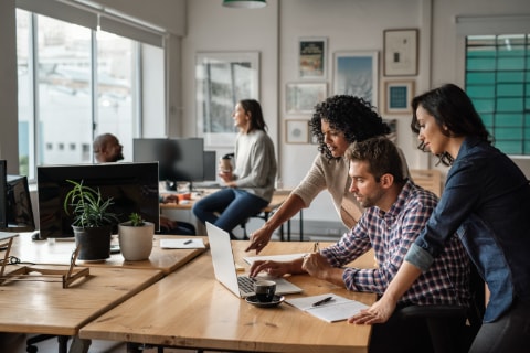 Photo of three people huddling around a computer in an office working on a creative endeavour.